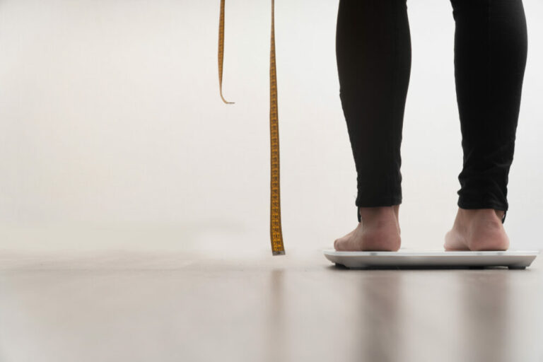 New FDA approved game changer in weight management - image of a white woman in black leggings standing on a scale and holding a measuring tape in a light, nondescript room